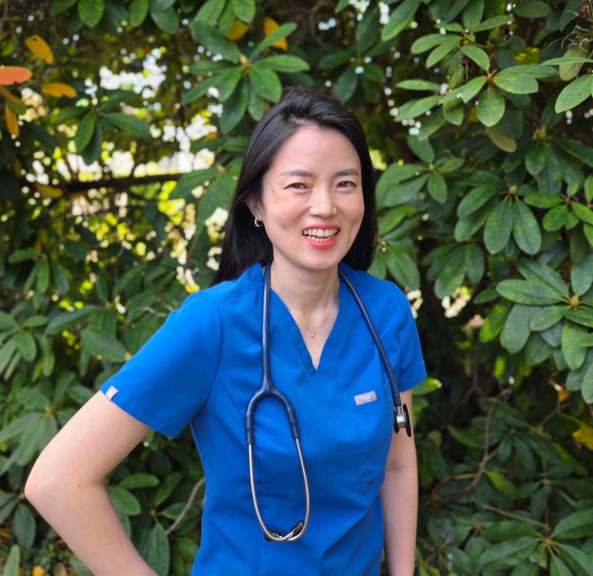 Miya Yi smiling in blue scrubs with a stethoscope, standing in front of a leafy rhododendron.